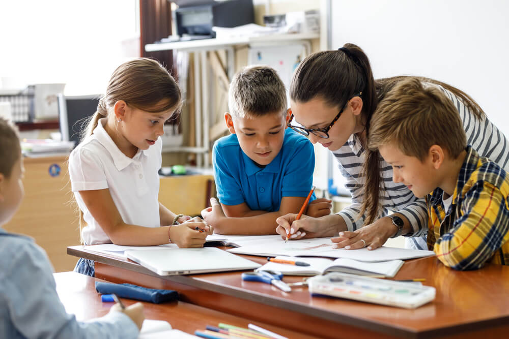 children writing on papers with school teacher in classroom