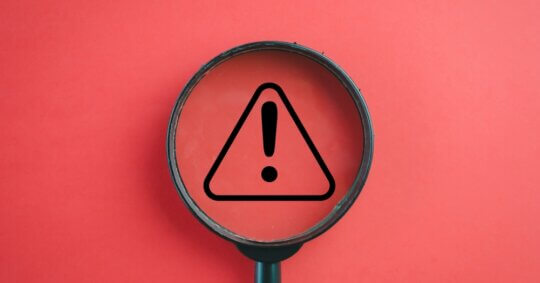 A magnifying glass covering a black warning icon over a red background.