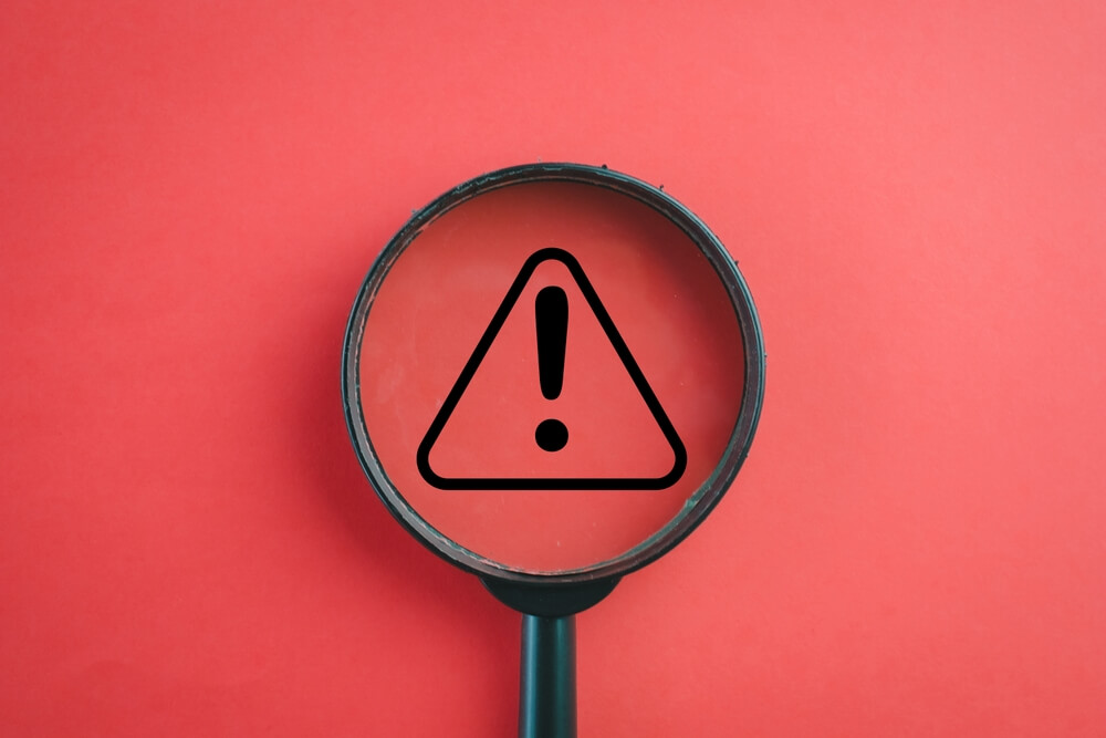 A magnifying glass covering a black warning icon over a red background.