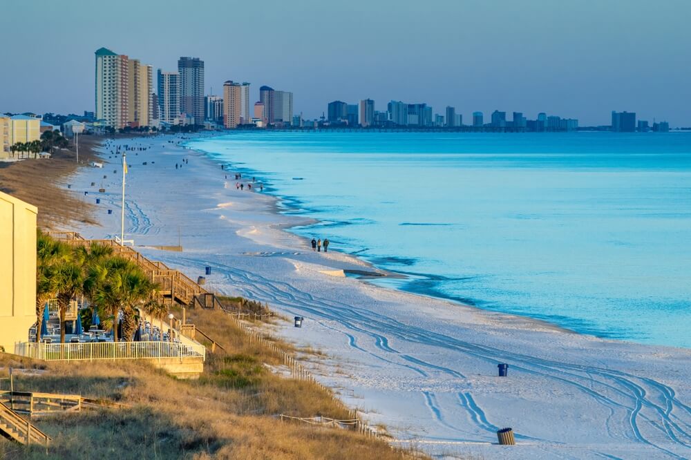 Photo of a landscape view of Panama City Beach in Florida with high-rise buildings, people walking along the shoreline, and soft ocean waves.