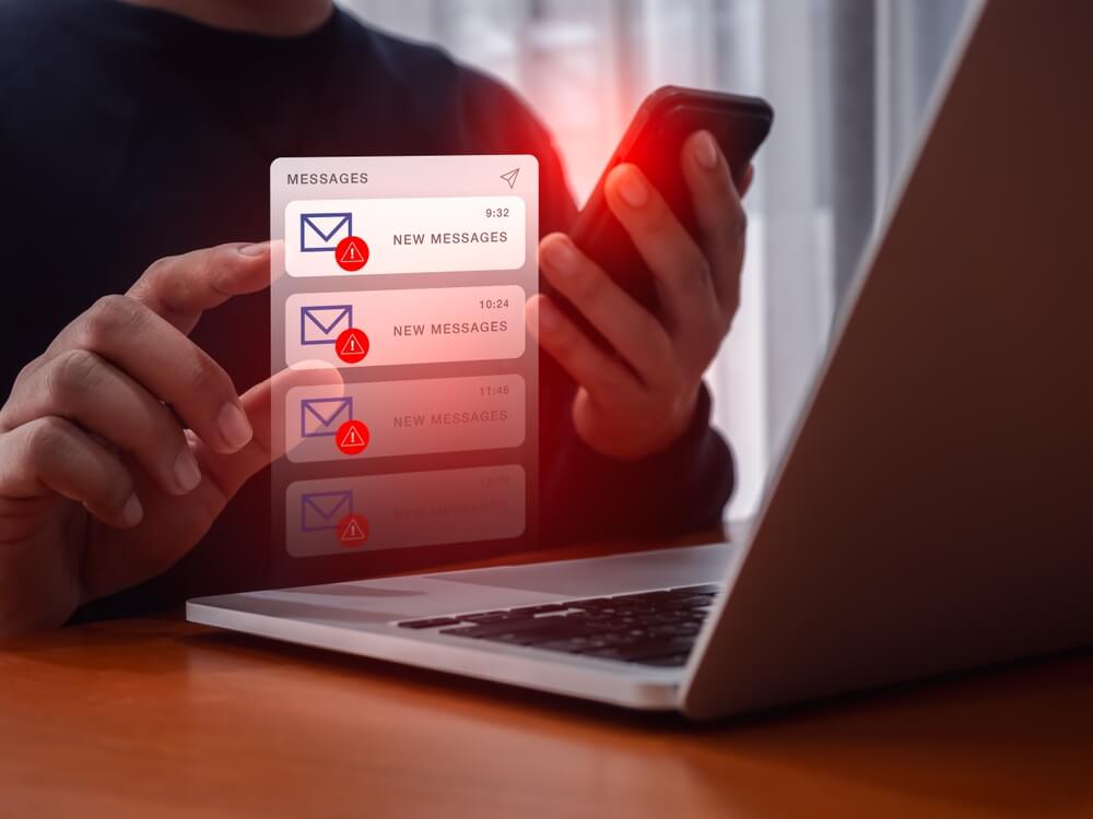 Photo of a person holding a smartphone and checking a laptop, with digital alerts and red warning icons indicating suspicious or potentially catfish emails and messages.